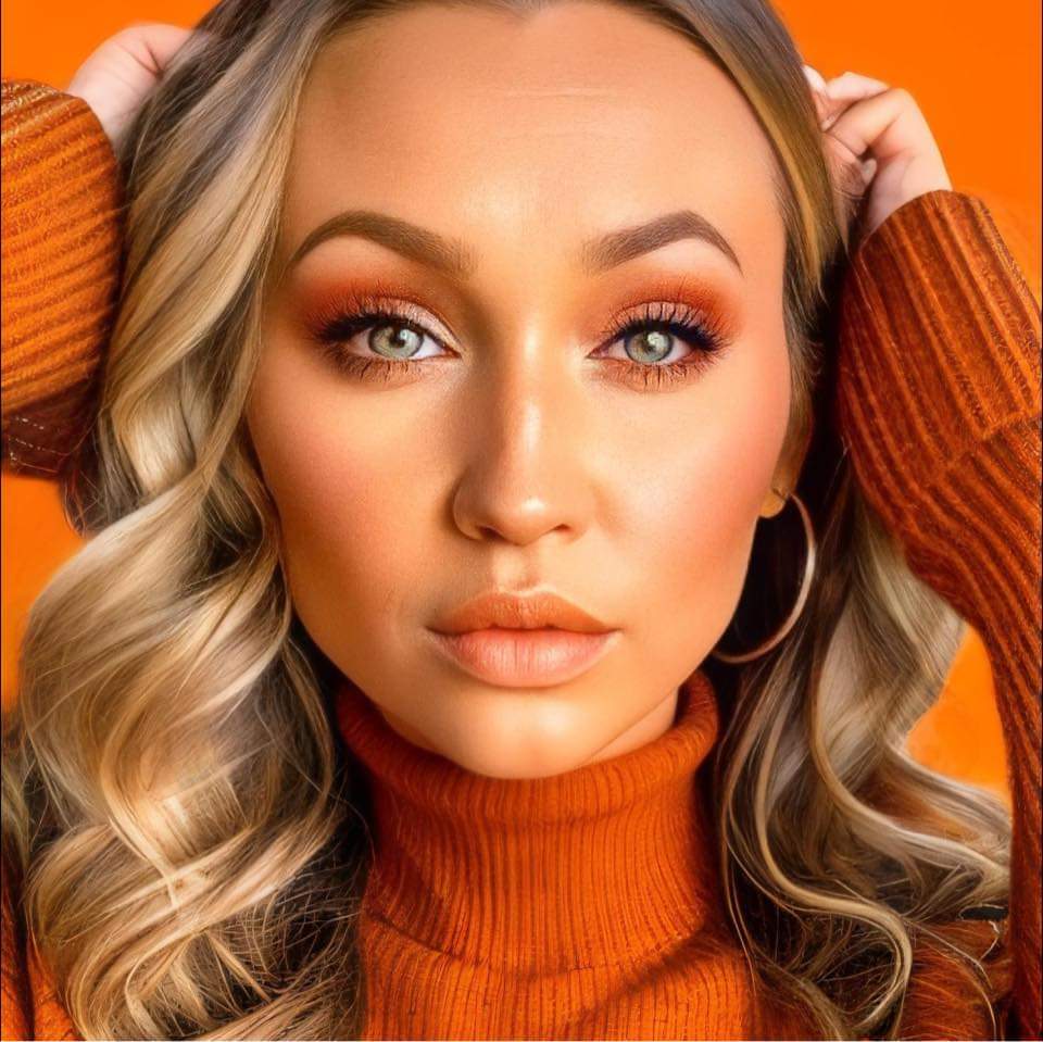 A woman in an orange sweater is posing for a photo wearing FACES by Brandi Special Occasion Makeup.